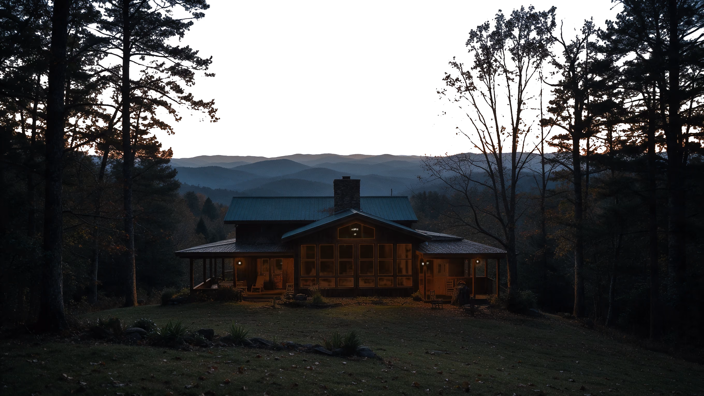 The Kalmia Woods cabin at dusk: dark board-and-batten walls, a green metal roof and a stone chimney, on a wooded rise above layered Blue Ridge mountains.